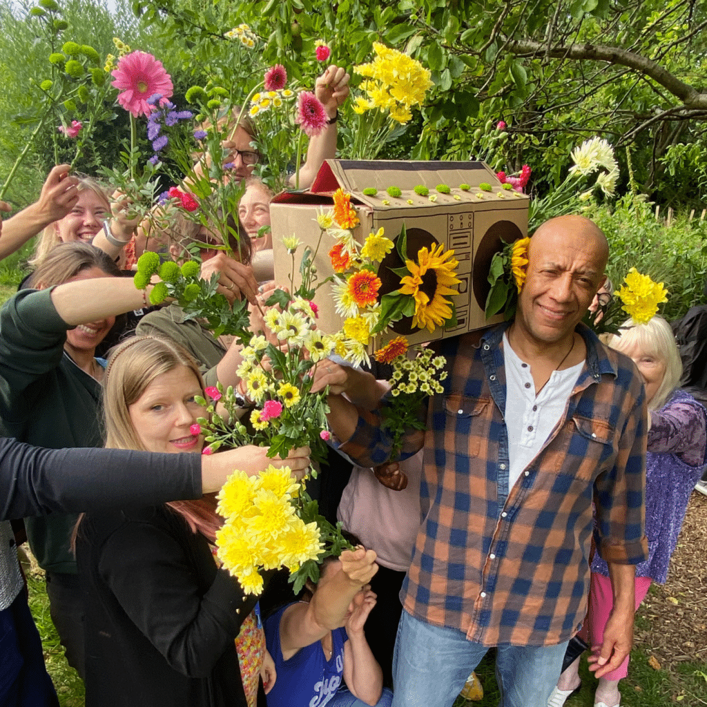 A man holds a boombox speaker made of cardboard, with a group of smiling people close behind him holding flowers in the air